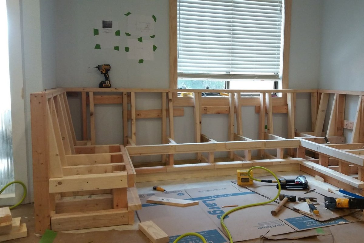 Wood-framed L-shaped bench seat built from 2x4 lumber under a window, with a drill and nail gun on the floor