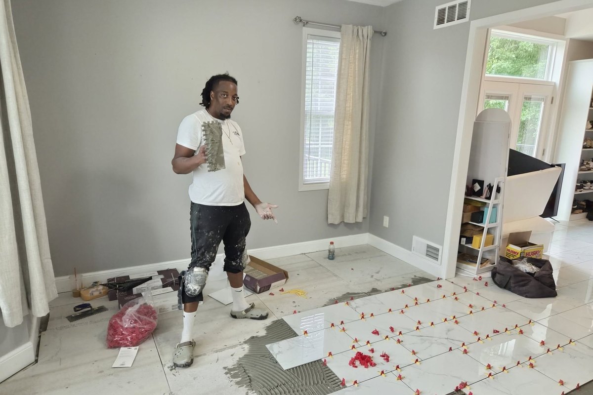 Tile setter in knee pads holding a notched trowel beside large white marble-look floor tile