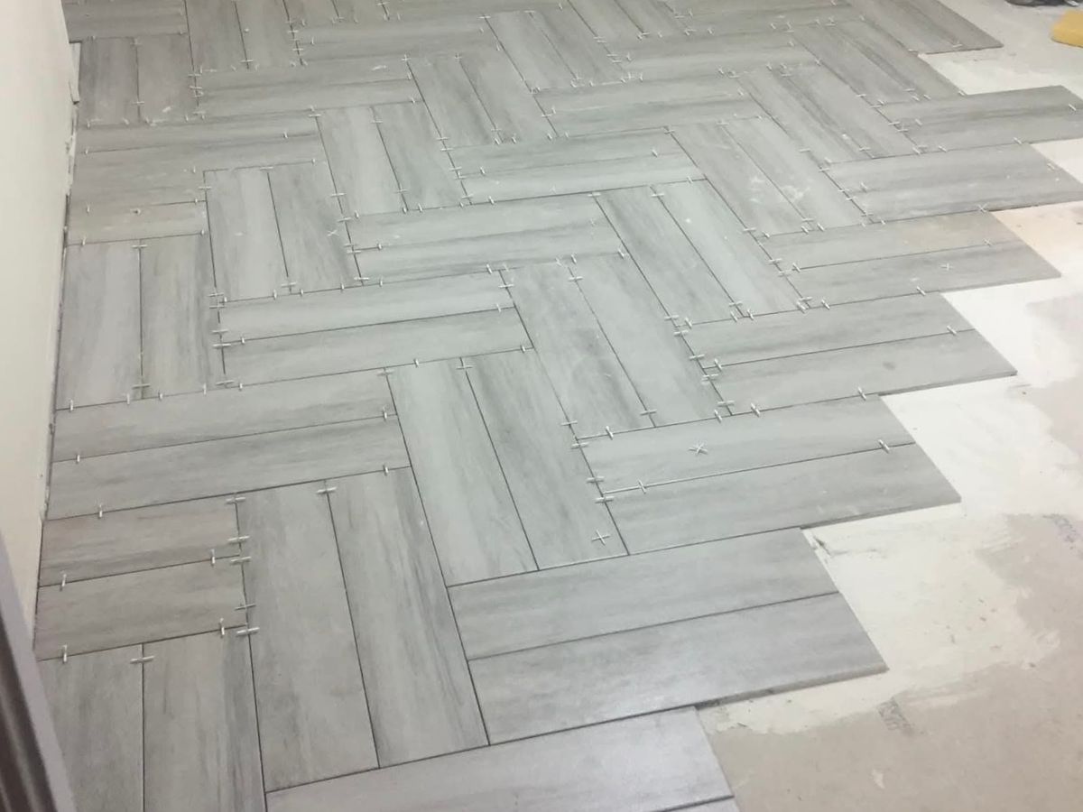 Gray wood-look rectangular porcelain tiles being laid in a herringbone pattern across a bedroom-size room