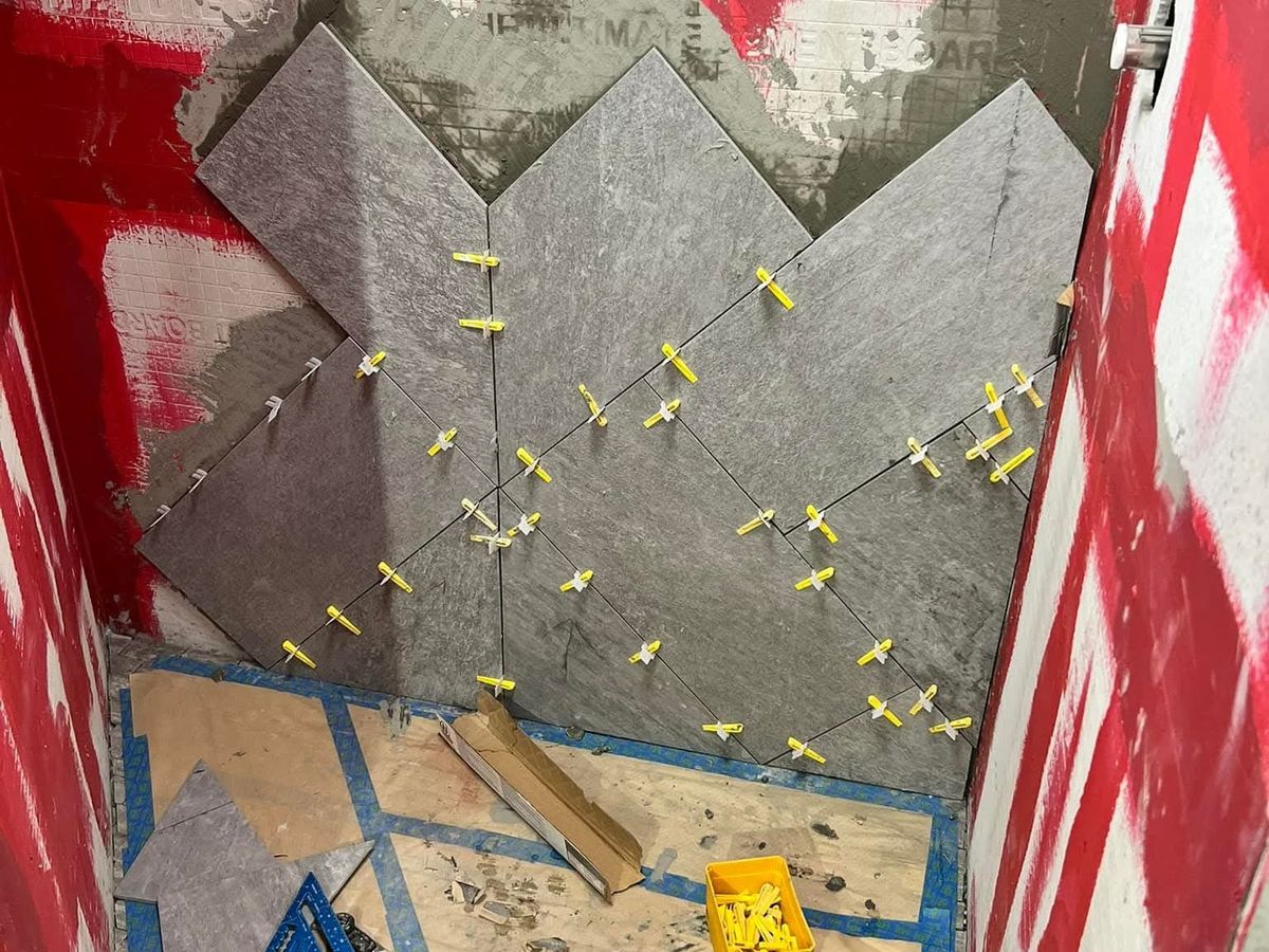 Chevron-pattern dark gray tile going up a shower back wall over red waterproofing, held by leveling clips