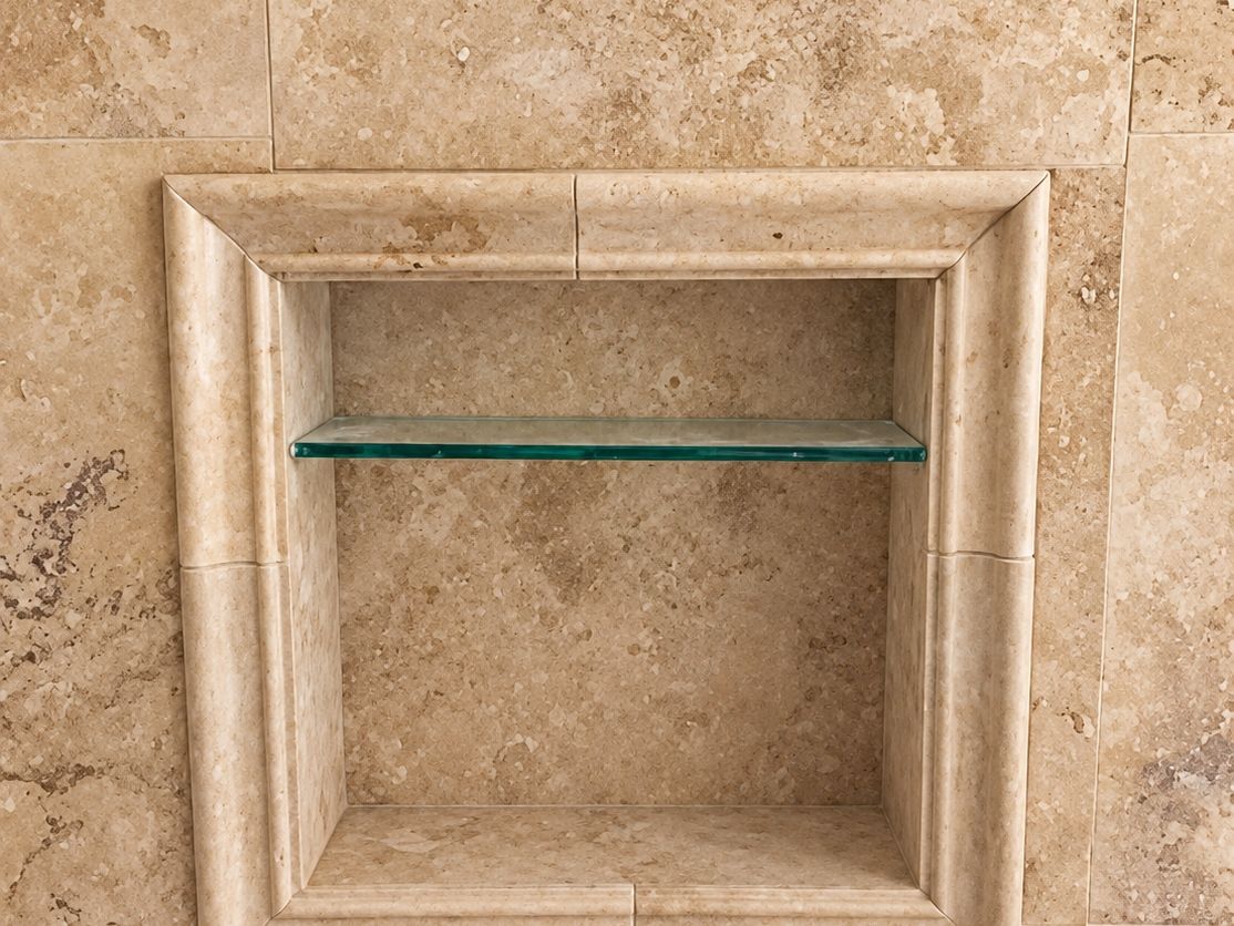 Close view of a finished shower niche framed with a carved travertine surround and a glass shelf