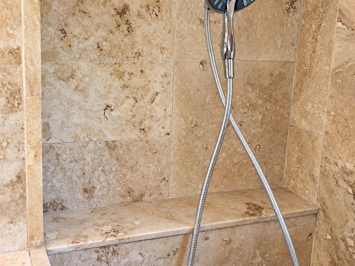Finished shower wall of large tan travertine tile with a chrome hand shower on a hose