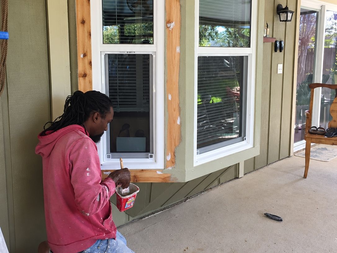 Worker in a pink hooded shirt kneeling to caulk the cedar frame of a new bay window with white sashes on green siding
