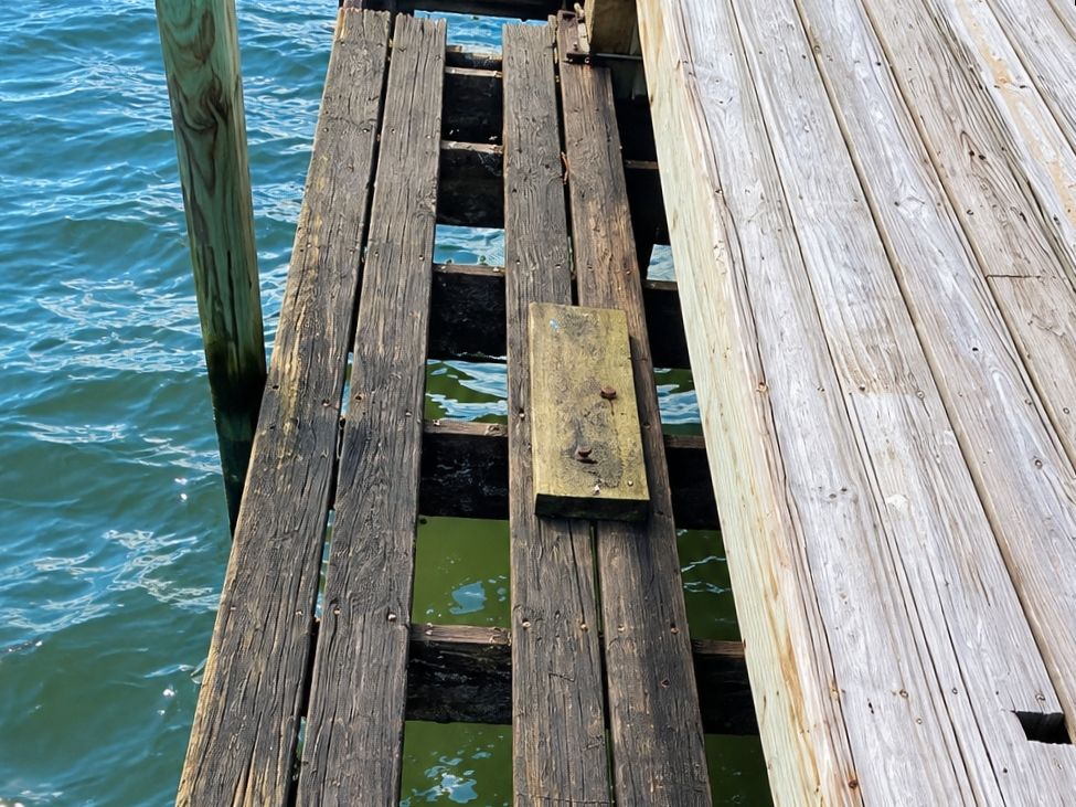 Weathered wooden dock boards with rotted and missing planks over green water beside one fresh light-colored plank
