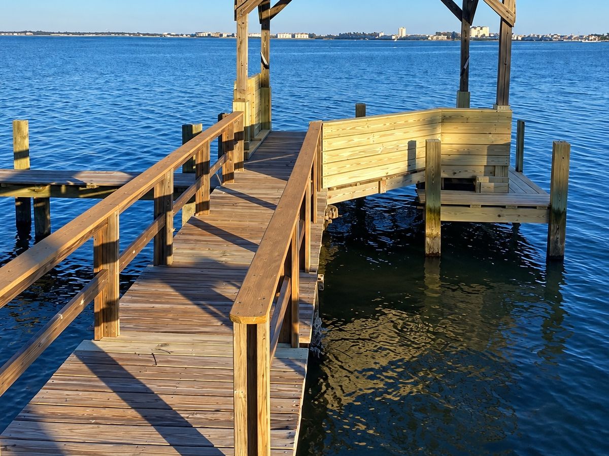 Wood boat dock walkway with railings leading over blue water to a platform with a pergola roof and bench seating