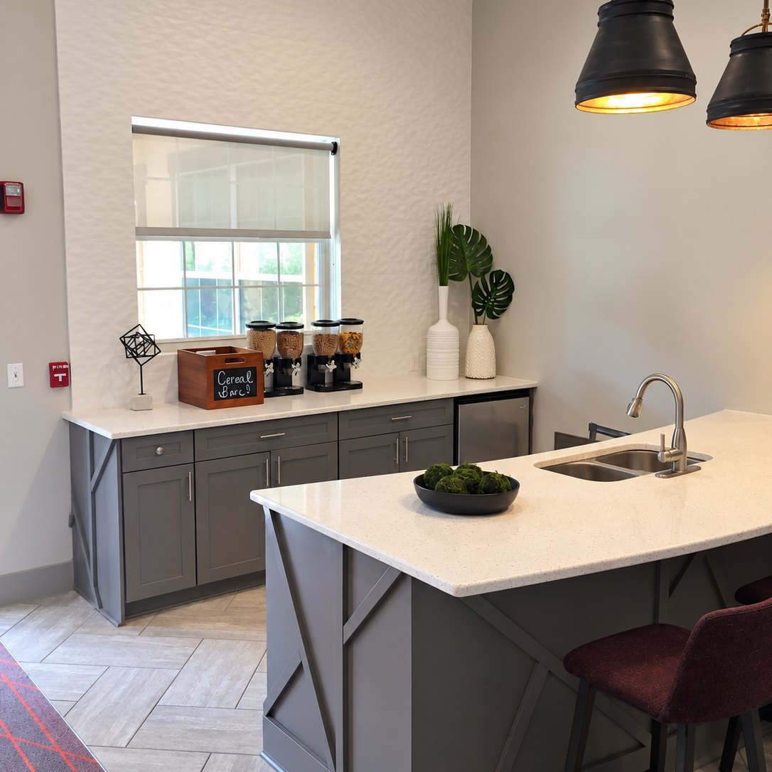 Break area kitchen with a textured white wall, gray cabinets, a white-topped angular-trim island, and black pendant lights